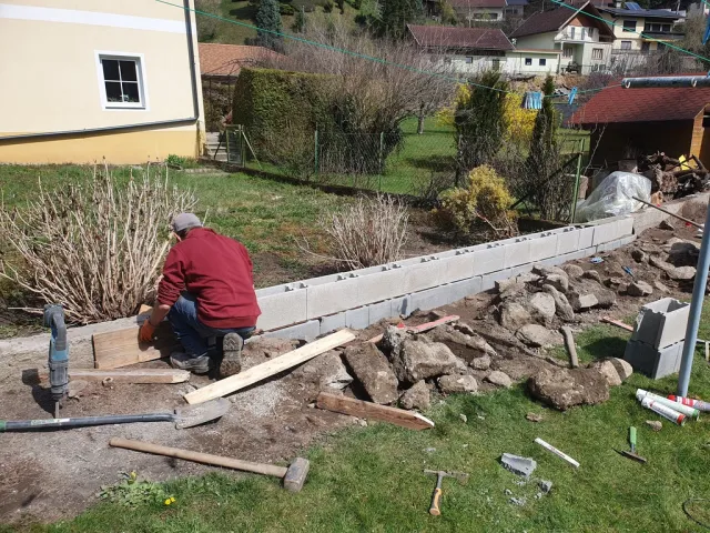 Ein Mann arbeitet im Garten und baut eine Mauer aus Betonsteinen. Werkzeuge und Baumaterialien liegen auf dem Rasen.