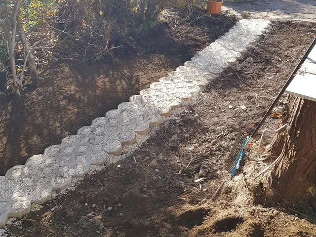 Ein Gartenweg aus grauen Pflastersteinen schlängelt sich durch einen Garten. Die Gartenarbeit ist im Gange, mit frischer Erde und einem Rechen in der Nähe.