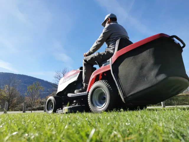 Ein Mann sitzt auf einem roten Rasenmäher und mäht den Rasen. Der Himmel ist blau und es gibt einige Bäume im Hintergrund.